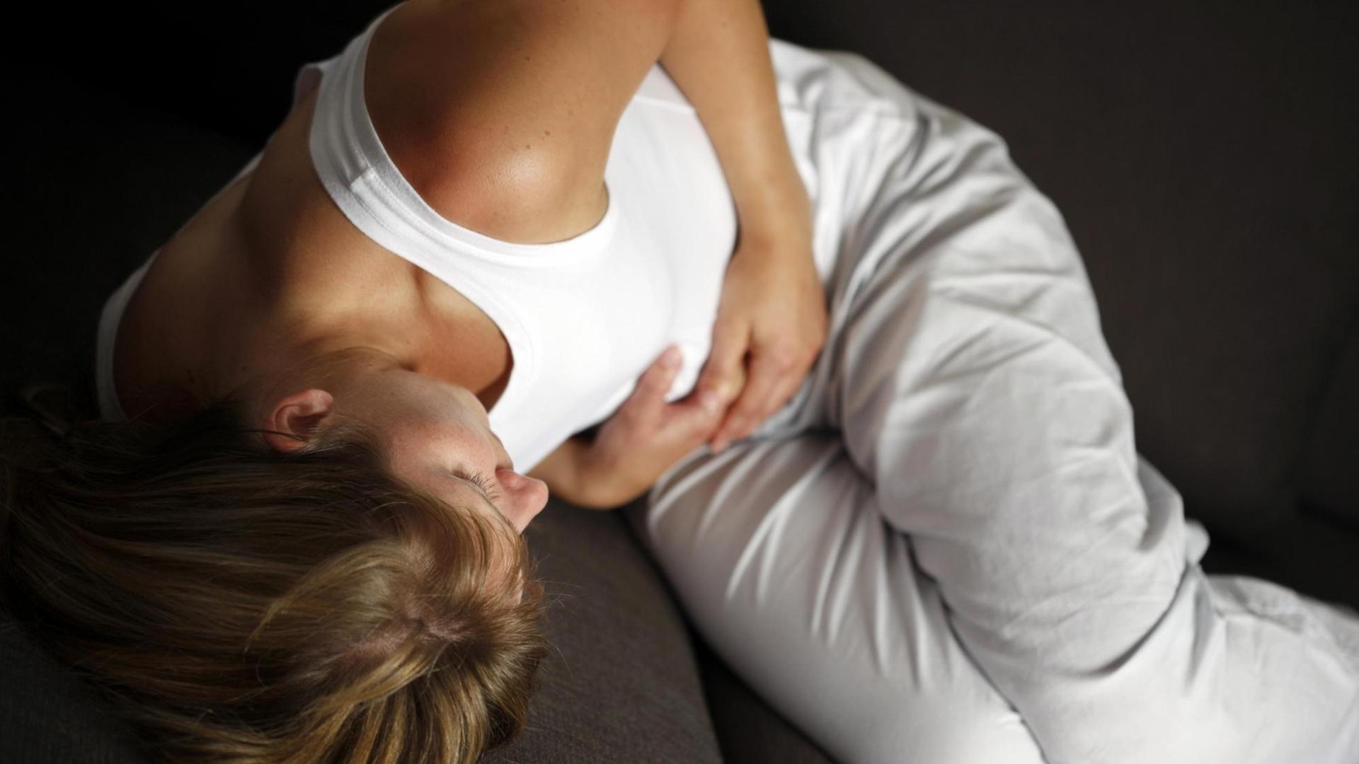 Symbolbild Leibschmerzen: Eine junge Frau liegt gekrümmt auf einem Sofa und hält sich die Hände auf die Magengegend.