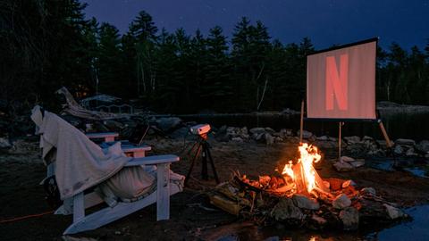 Eine Leinwand, ein Projektor, an einem See mit Lagerfeuer unter dem Sternenhimmel.