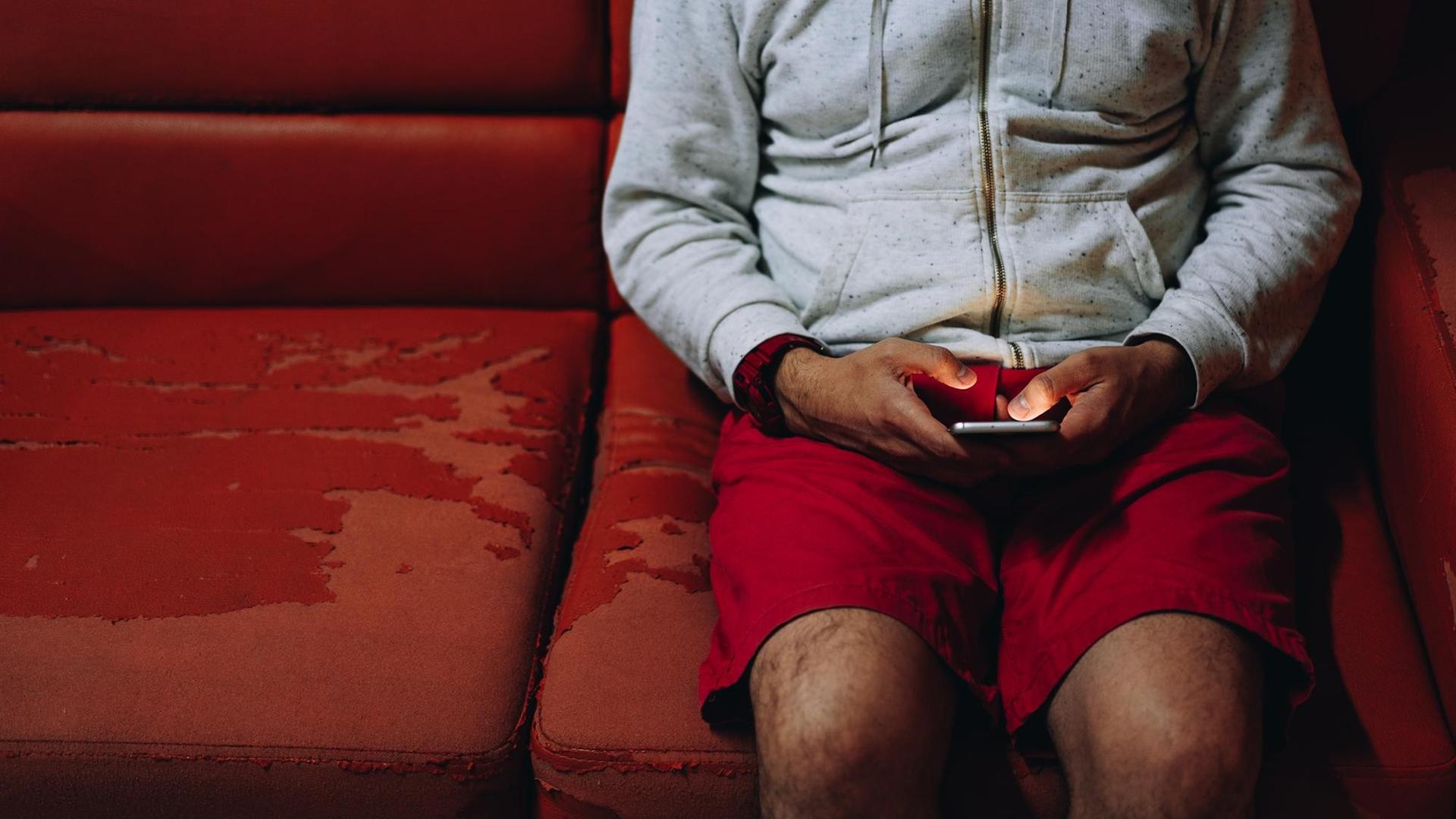 Ein Mann sitzt auf einer alten roten Couch und schaut auf sein Handy.