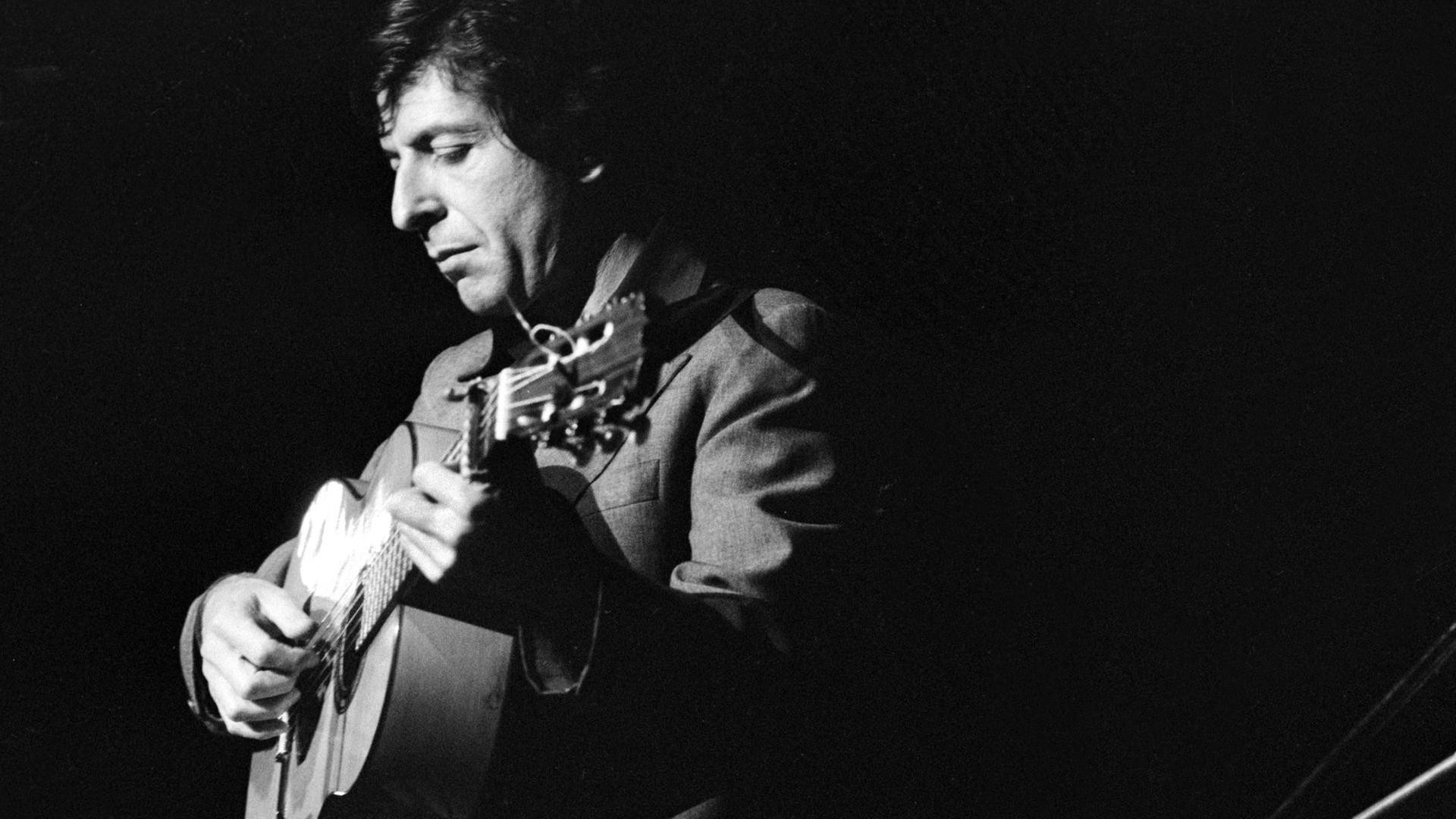 Schwarzweißfoto von Leonard Cohen, der nachdenklich und Gitarre spielend auf einer Bühne steht