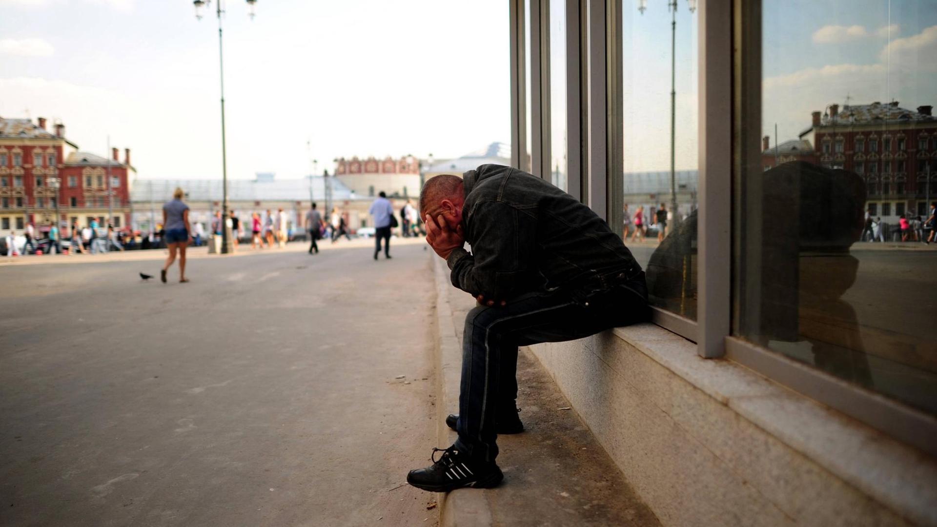 Ein junger Mann sitzt sitzt an ein Schaufenster in Moskau gelehnt und stützt das Gesicht in die Hände