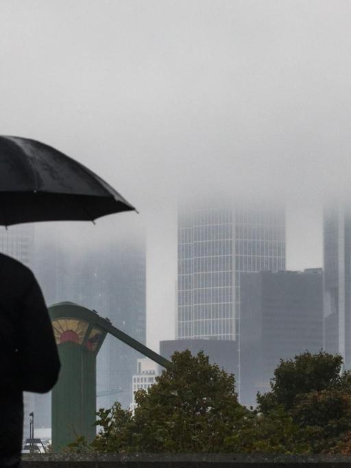 Ein Mann geht am 24.10.2016 in Frankfurt am Main (Hessen) mit aufgespanntem Schirm an der regnerischen Kulisse der Stadt vorbei. Die Spitzen der Hochhäuser verschwinden im Nebel.