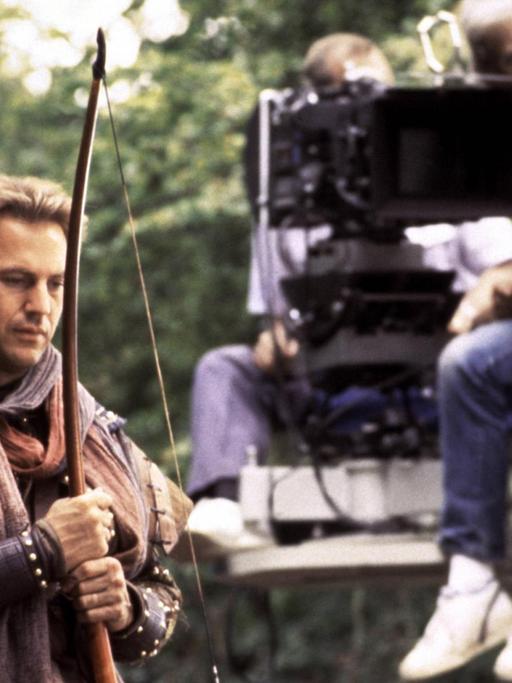 Kevin Costner steht im Robin-Hood-Kostüm samt Bogen vor einem Kamerakran