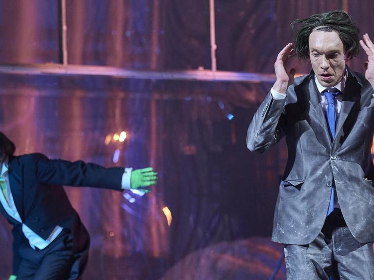 Szenenfoto: Ein Mann im Anzug und mit zerzausten Haaren hebt die Hände Richtung Kopf. Neben ihm steht ein Mann, der aussieht wie die Comic-Figur "Joker". Verwüstung auf der Bühne.
