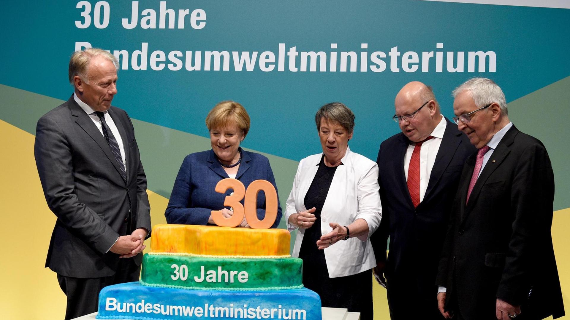 Die ehemaligen Umweltminister Jürgen Trittin und Kanzlerin Angela Merkel stehen mit der amtierenden Umweltministerin Barbara Hendricks (SPD), Kanzleramtschef Peter Altmaier (CDU) und Klaus Töpfer (CDU) vor einer Torte, auf der zu lesen ist: 30 Jahre Bundesumweltministerium.