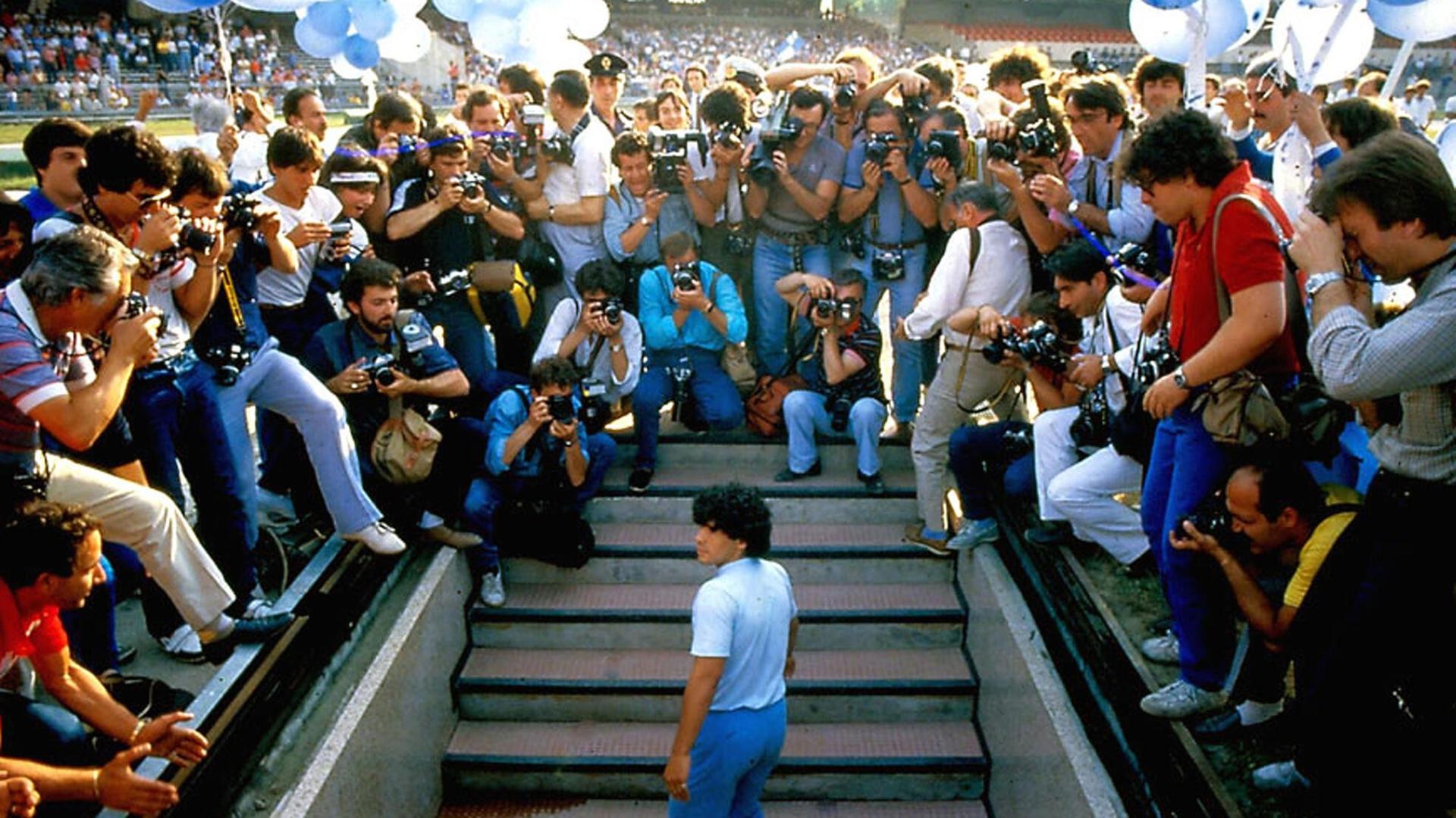 Im Still aus "Diego Maradona" steht der Fußballspieler von Fotografen umringt auf einer Treppe.