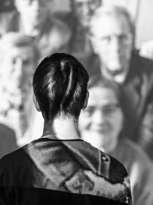 Eine Person steht mit dem Gesicht zur Wand vor einer Videoinstallation auf der viele ältere Menschen zu sehen sind.
