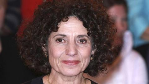 Eine Frau mit dunklen Locken lächelt und schaut nach links knapp neben der Kamera vorbei.