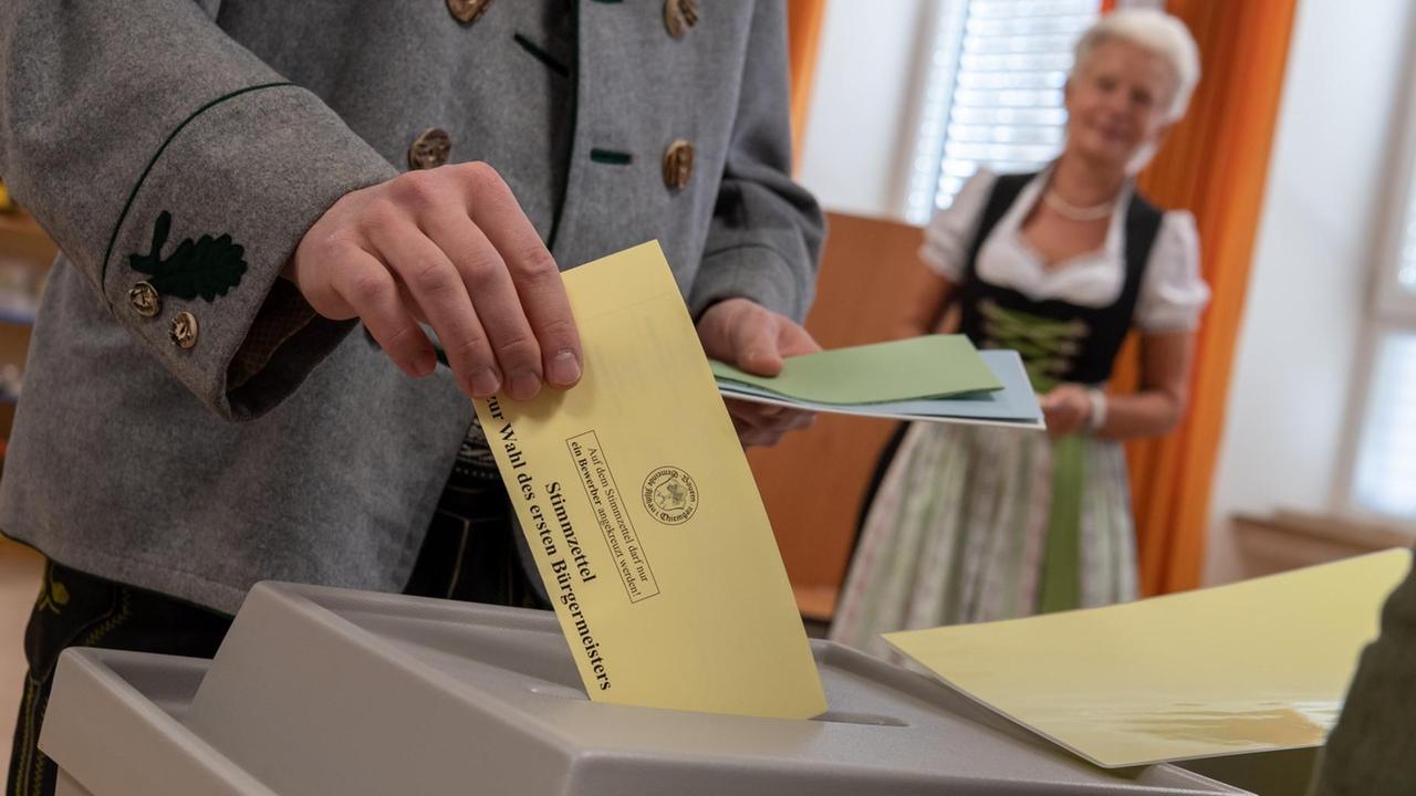 Ein Mann wirft einen Wahlzettel in die Urne. Im Hintergrund steht eine Frau. Beide tragen Tracht.