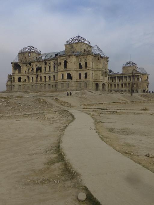 Ruine des Darul-Aman-Palastes: ein rechteckiges pompöses und zerbombtes Gebäude aus der Ferne, eine einsame Straße führt nach dort