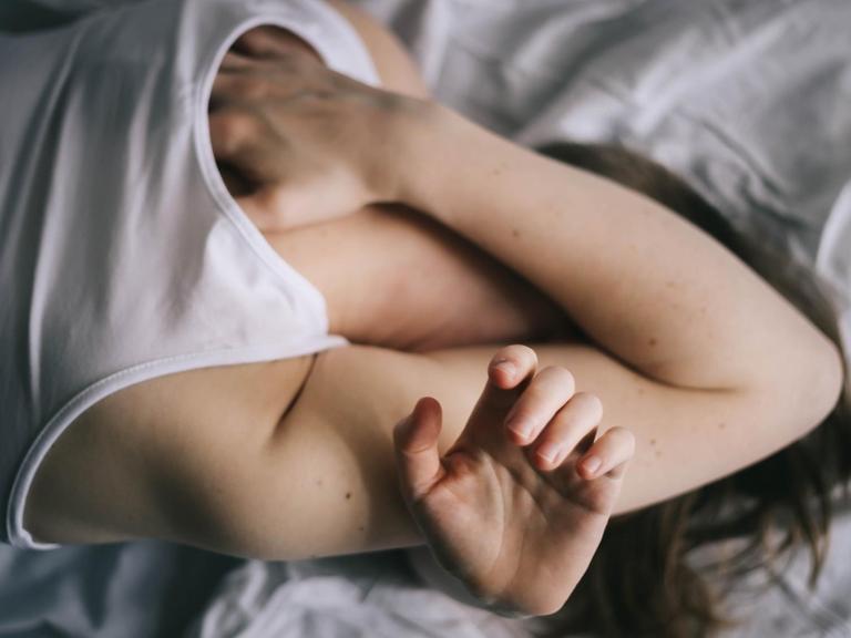 Eine Frau liegt im Bett und verbirgt das Gesicht unter ihren Armen.
