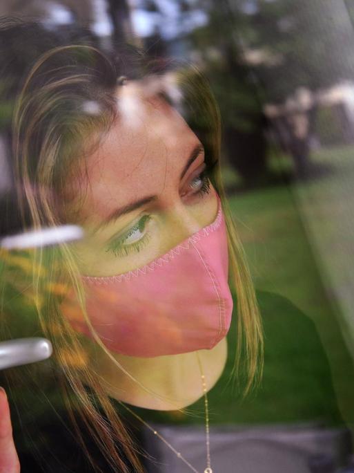 Eine junge Frau mit Schutzmaske steht am Fenster in einem Zimmer und blickt hinaus.