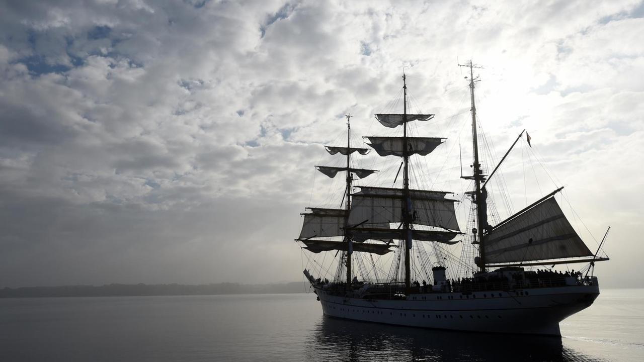Das Bild zeigt das Schiff im Zwielicht unter wolkigem Himmel im grauen Meer. Die Segel werden gerade gesetzt.