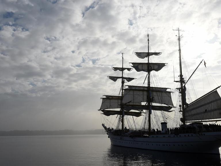 Das Bild zeigt das Schiff im Zwielicht unter wolkigem Himmel im grauen Meer. Die Segel werden gerade gesetzt.