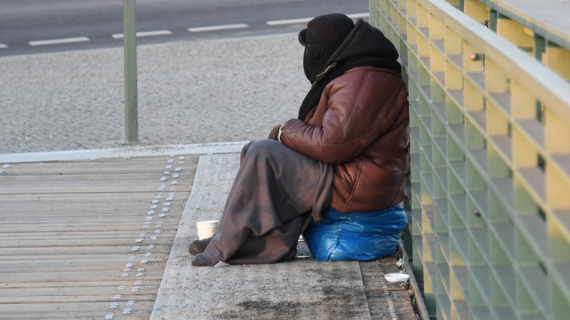 Ein Bettler, eingemummt in eine dicke Jacke,Pelzmuetze und Decke, sitzt bei bissiger Kaelte auf der Brücke vor dem Hauptbahnhof in Berlin.