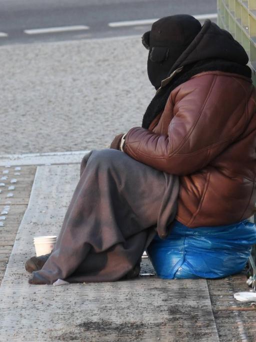 Ein Bettler, eingemummt in eine dicke Jacke,Pelzmuetze und Decke, sitzt bei bissiger Kaelte auf der Brücke vor dem Hauptbahnhof in Berlin.