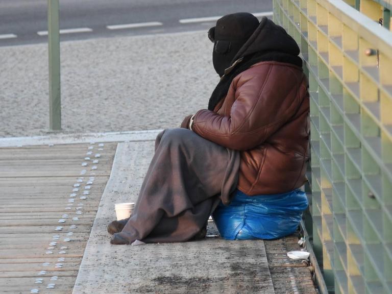 Ein Bettler, eingemummt in eine dicke Jacke,Pelzmuetze und Decke, sitzt bei bissiger Kaelte auf der Brücke vor dem Hauptbahnhof in Berlin.