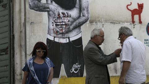 Menschen in Athen stehen vor einem Graffiti, das einen Mann zeigt, der ein Euro-Zeichen zerdrückt.