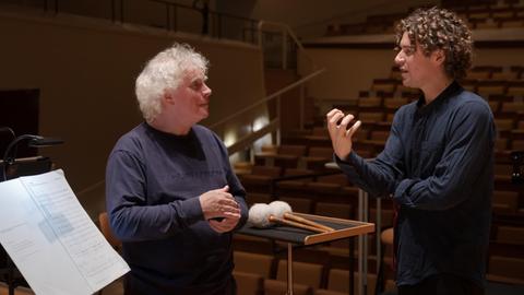Neben einem Notenpult (v.l.) diskutieren ein älterer grauhaariger Mann mit einem jüngeren Mann mit gelocktem Haar in einem leeren Konzertsaal.