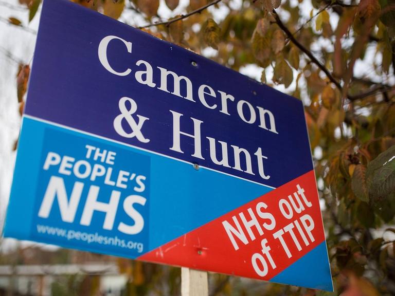 Ein Plakat an einem Stock wird in die Luft gehalten, darauf die Aufschrift: "Cameron & Hunt - The People's NHS - NHS out of TTIP"