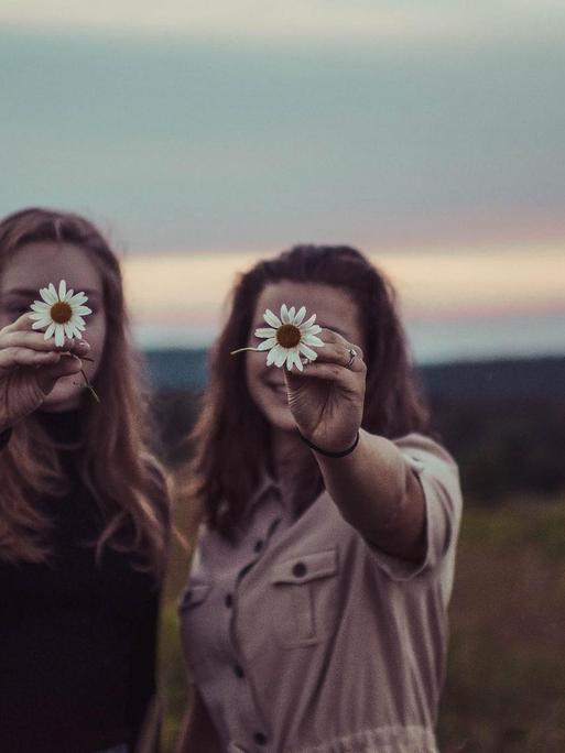Zwei Frauen stehen nebeneinander und halten sich eine Blume vor das Gesicht.