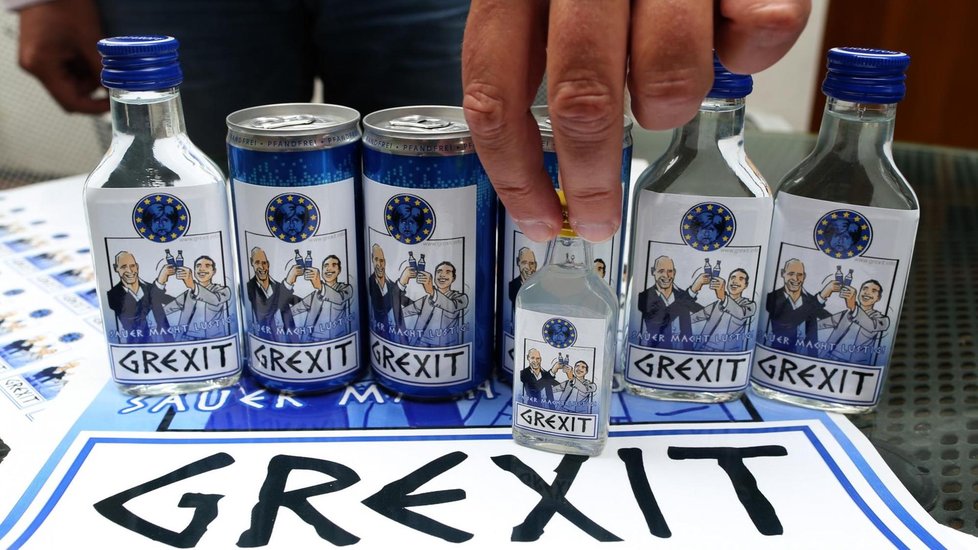 Auf einem Tisch stehen mehrere Flaschen und Dosen, auf den Etiketten stoßen Politiker an, darunter das Wort "Grexit".