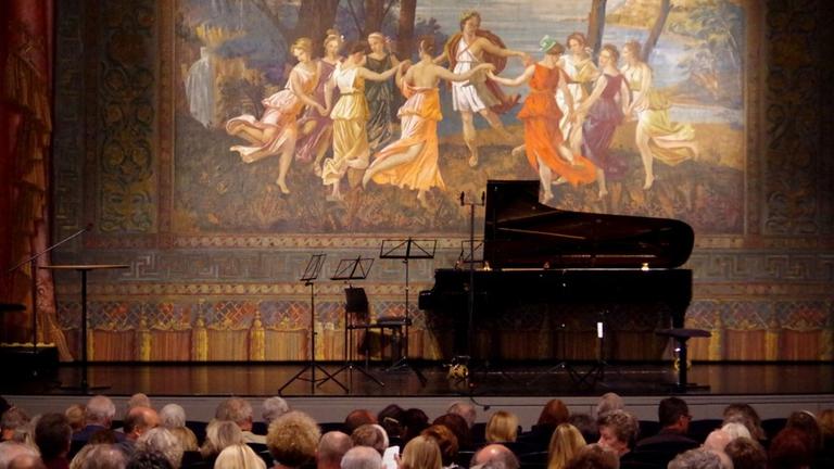 Auf der Theaterbühne von Kempten mit dekorativem Vorhang steht ein aufgeklappter Flügel und Notenständer. Das Publikum wartet auf die Musiker.
