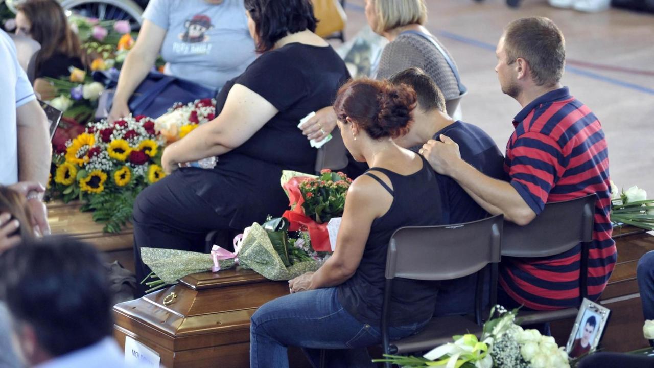 Trauernde Menschen sitzen mit gesenkten Köpfen vor Särgen.