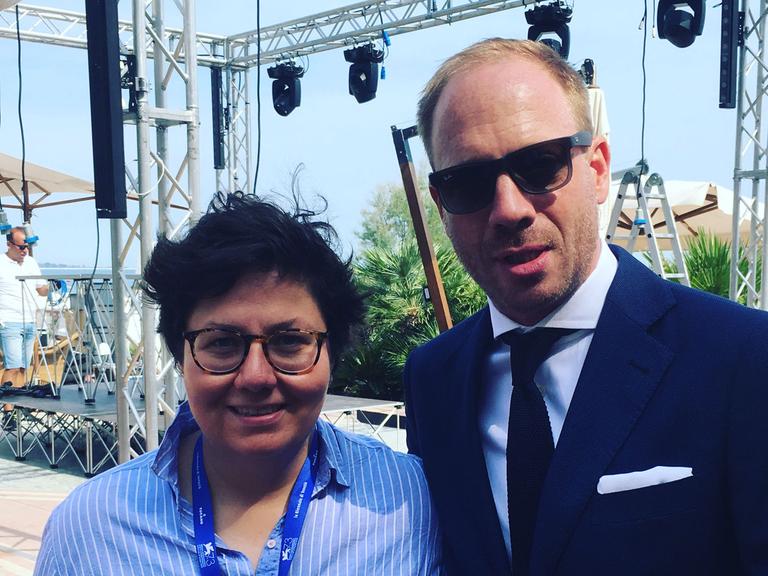Schauspieler Johann von Bülow und die Autorin Anna Wollner bei den Filmfestspielen in Venedig 2016. Sie hat ihn einen Tag lang auf dem Lido begleitet. Beide lächeln fröhlich in die Kamera. Johann von Bülow trägt eine Sonnenbrille.