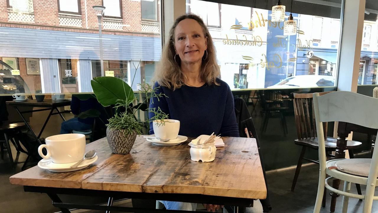 Eine blonde Frau sitzt in einem Café an einem hohen Tisch, auf dem zwei Kaffeetassen stehen.