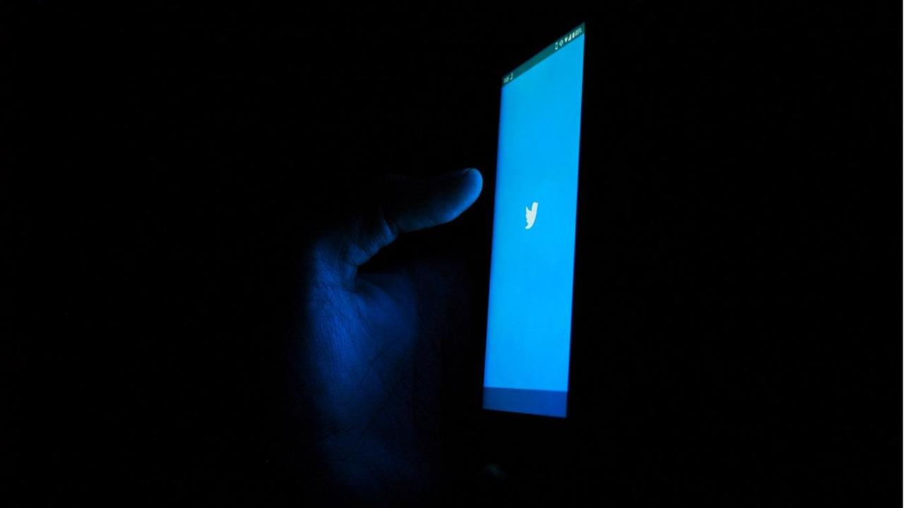 Eine Hand hält ein Smartphone mit blauem Twitterlogo auf dem Display in der Dunkelheit.