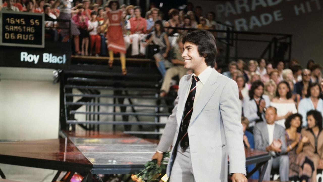 Ein Farbbild zeigt den  Schlagersänger Roy Black in  weißgrauem Anzug und 70er-Jahre-Frisur bei einem Auftritt in der ZDF-Hitparade
