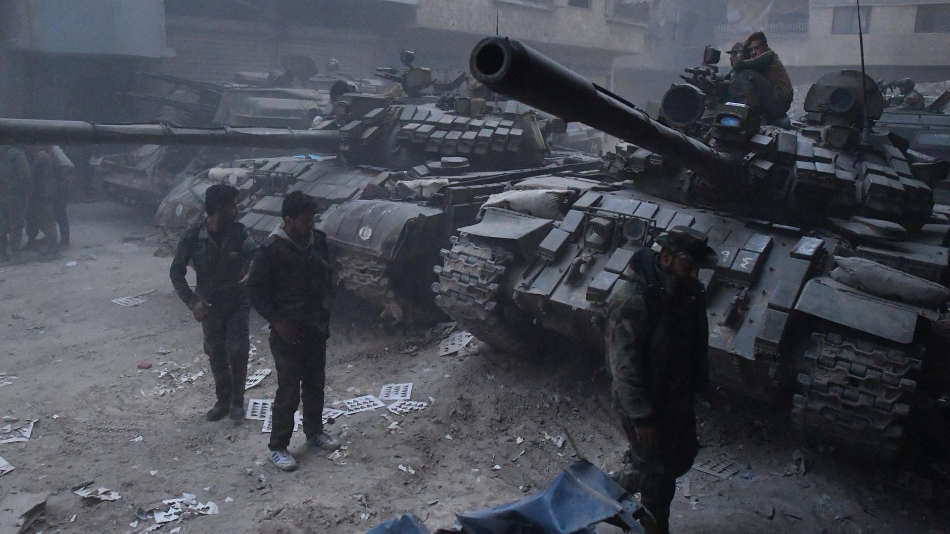 Zwei Panzer stehen auf einer nicht asphaltierten Straße, im Hintergrund Nebel und Wohnhäuser, vor den Panzern gehen drei Männer.