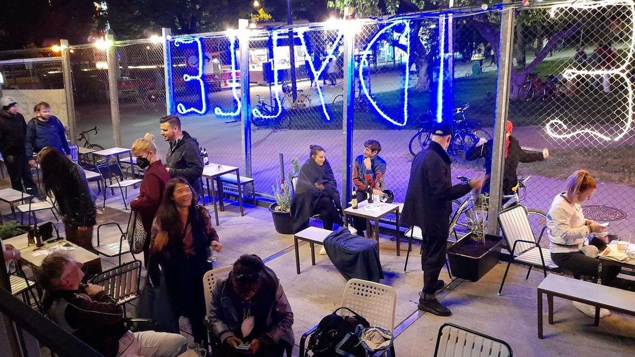 Im Hof des Clubs sitzen viele Mensche in Jacken und Decken gehüllt an Tischen und nehmen Getränke zu sich. Am Zaun steht in Neonbuchstaben "Idylle" geschrieben.