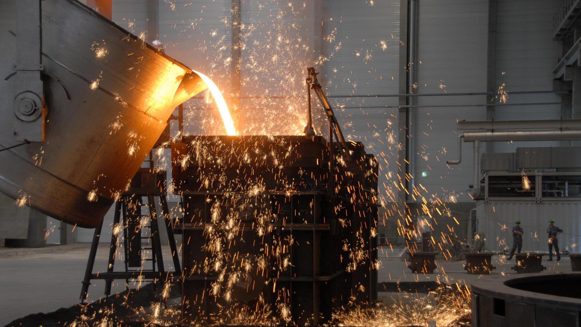 Flüssiges Eisen wird von einem Behälter in eine Form geegossen, dabei sprühen Funken. Im Hintergrund stehen Arbeiter in der Eisengießerei Torgelow