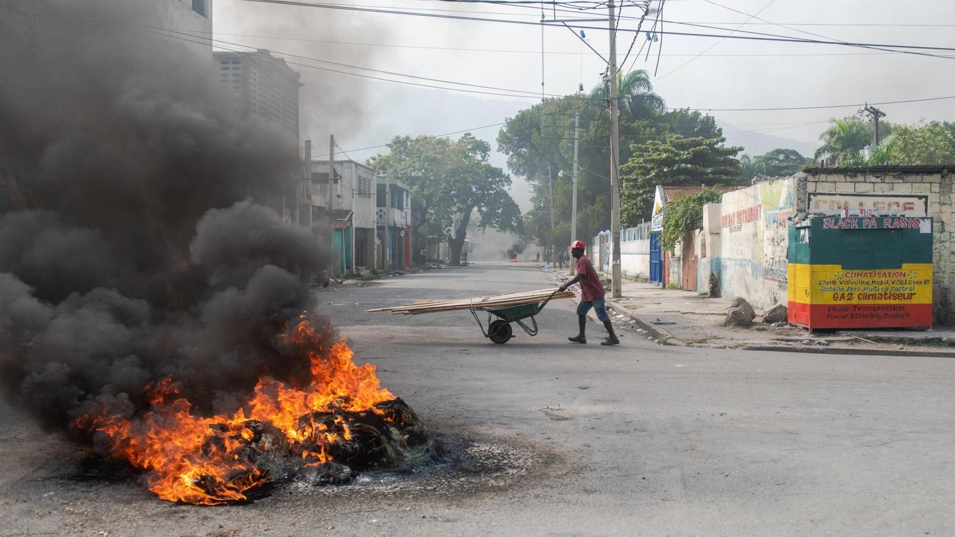 Eine brennende Barrikade in Haitis Hauptstadt Port-au-Prince. Dahinter geht eine Person mit einer Schubkarre die Straße entlang.