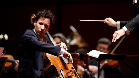 Ein junger Cellist sitzt vor einem Orchester und spielt konzentriert sein Instrument.