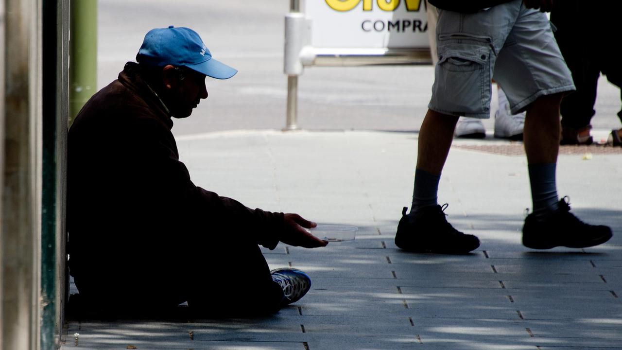 Ein Bettler mit einem blauen Käppi sitzt auf dem Boden in einer Fußgängerzone und streckt die rechte Hand aus.