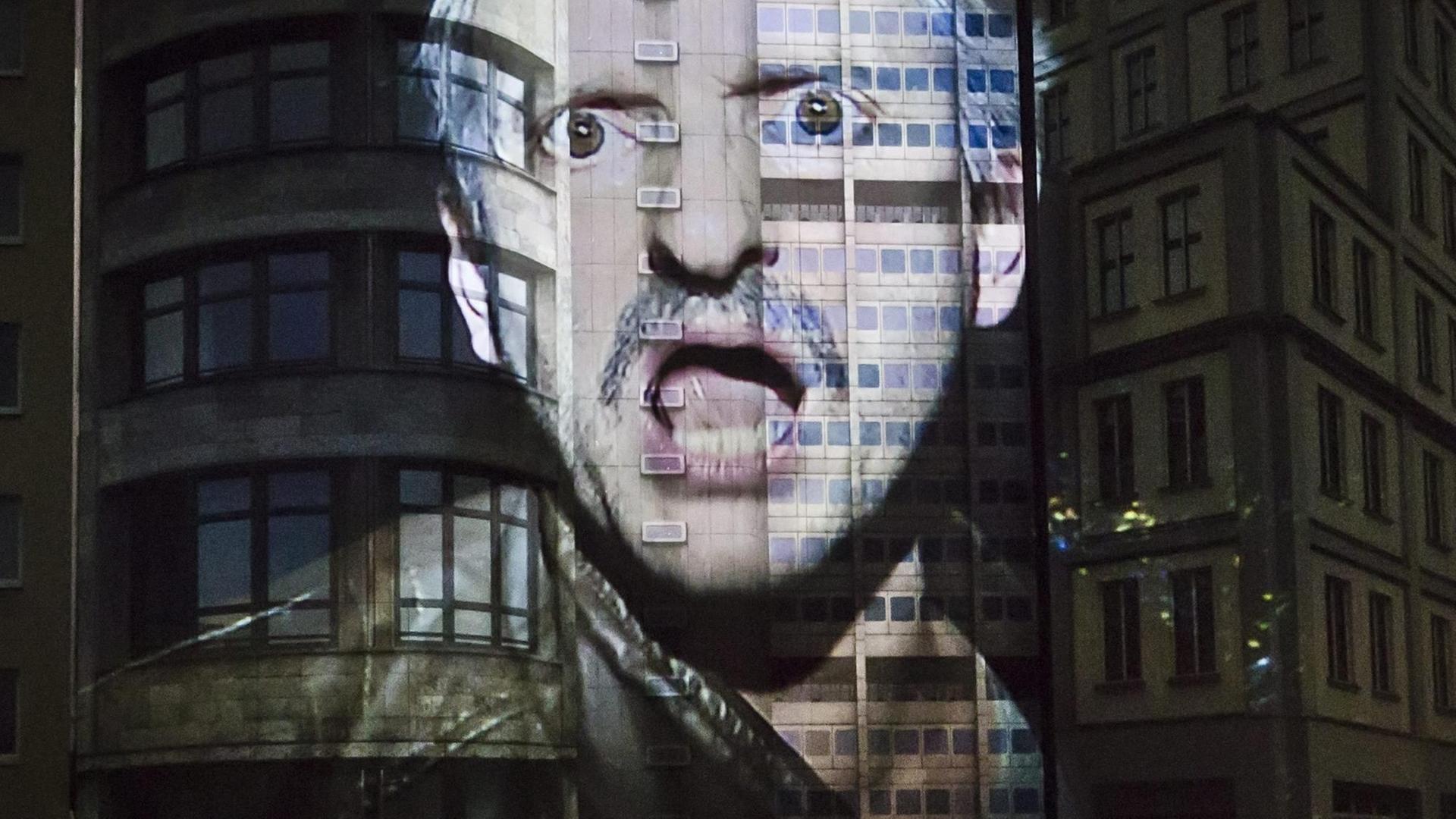In der Inszenierung wird das Gesicht von Milan Peschel mit empörtem Ausdruck an eine Gebäudefassade projiziert.