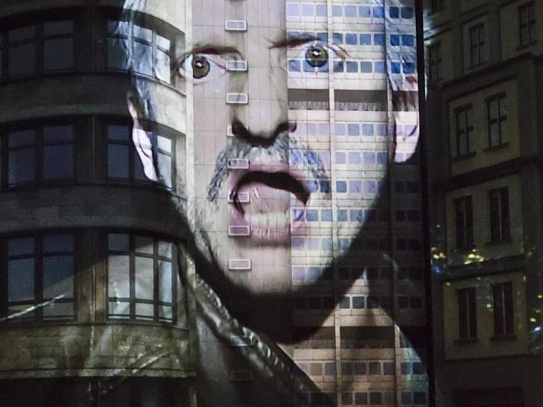 In der Inszenierung wird das Gesicht von Milan Peschel mit empörtem Ausdruck an eine Gebäudefassade projiziert.