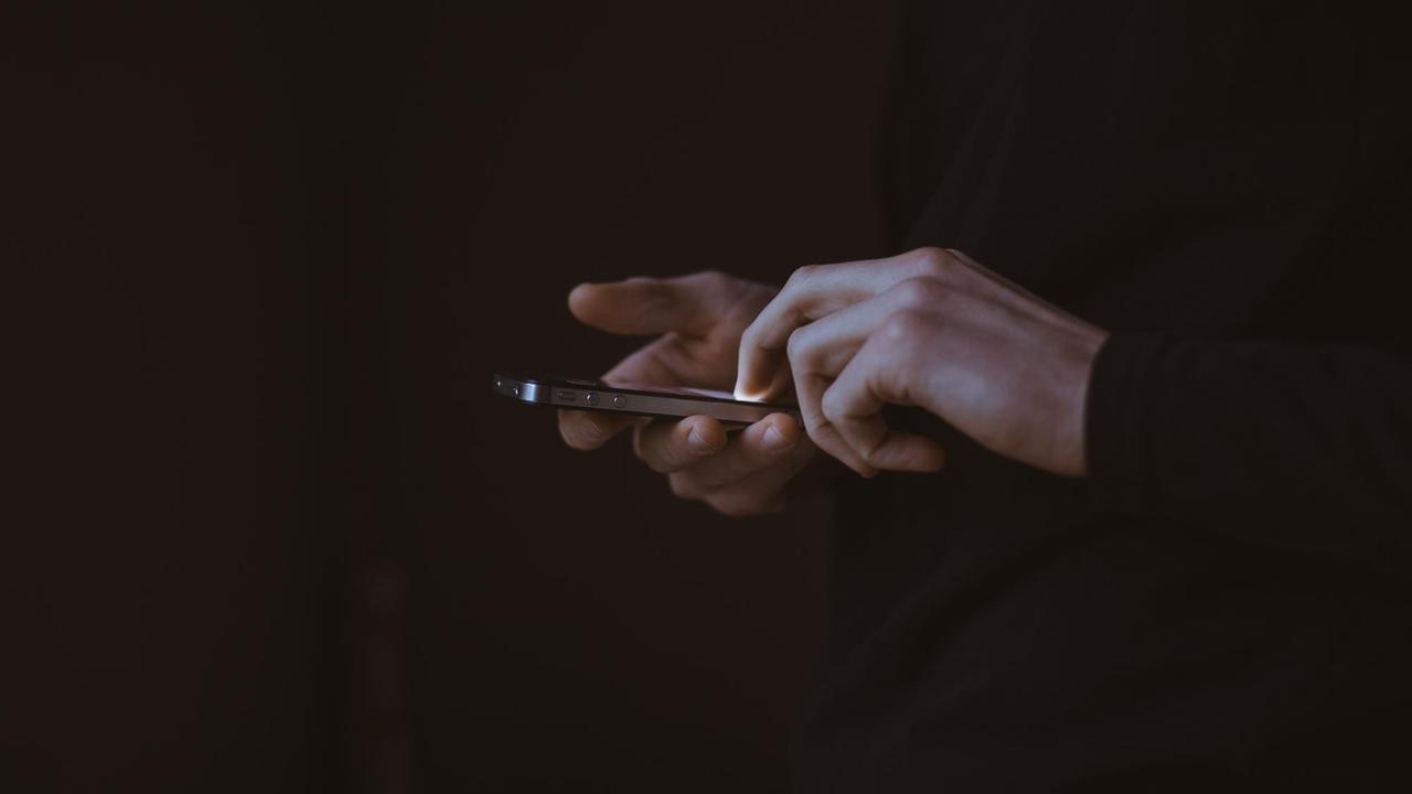 Das Bild zeigt zwei helle Hände vor sehr dunklem Hintergrund, die mit den Fingerspitzen ein leuchtendes Smartphone-Display bedienen.