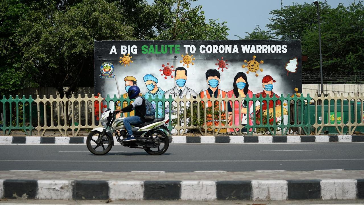 In Indiens Hauptstadt Neu Delhi würdigt ein Plakat den Kampf vieler Menschen gegen die Corona-Pandemie. Auf dem Plakat sind u.a. ein Arzt, eine Pflegerin und ein Polizist zu sehen. Darunter steht: "A big salute to corona warriors" ("Ein Gruß an die Corona-Kämpfer"). Ein Motorradfahrer fährt daran vorbei.