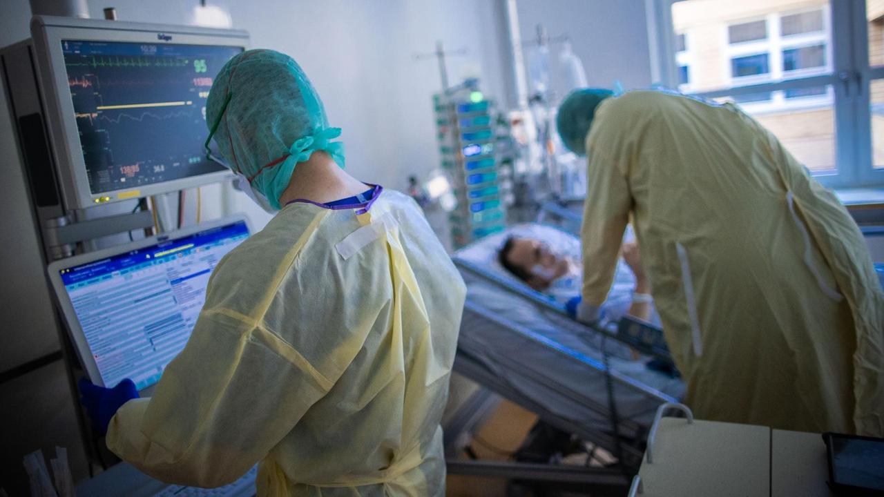 Zwei Krankenpfleger im Zimmer eines Patienten. Einer steht an einem Monitor, der andere beugt sich über das Bett. Beide tragen Kittel und Masken.