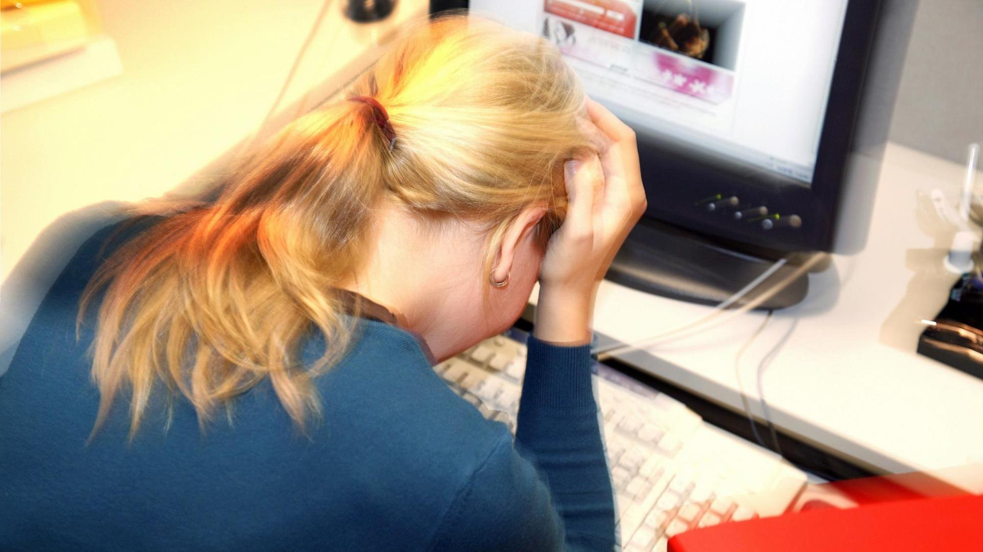 Eine Frau sitzt an einem Schreibtisch vor einem Computer, den Kopf auf die Hände gestützt.