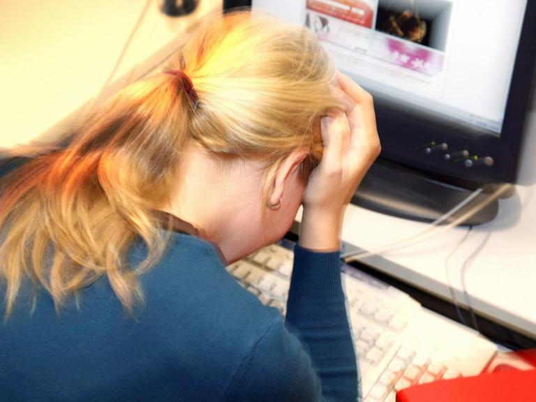 Eine Frau sitzt an einem Schreibtisch vor einem Computer, den Kopf auf die Hände gestützt.