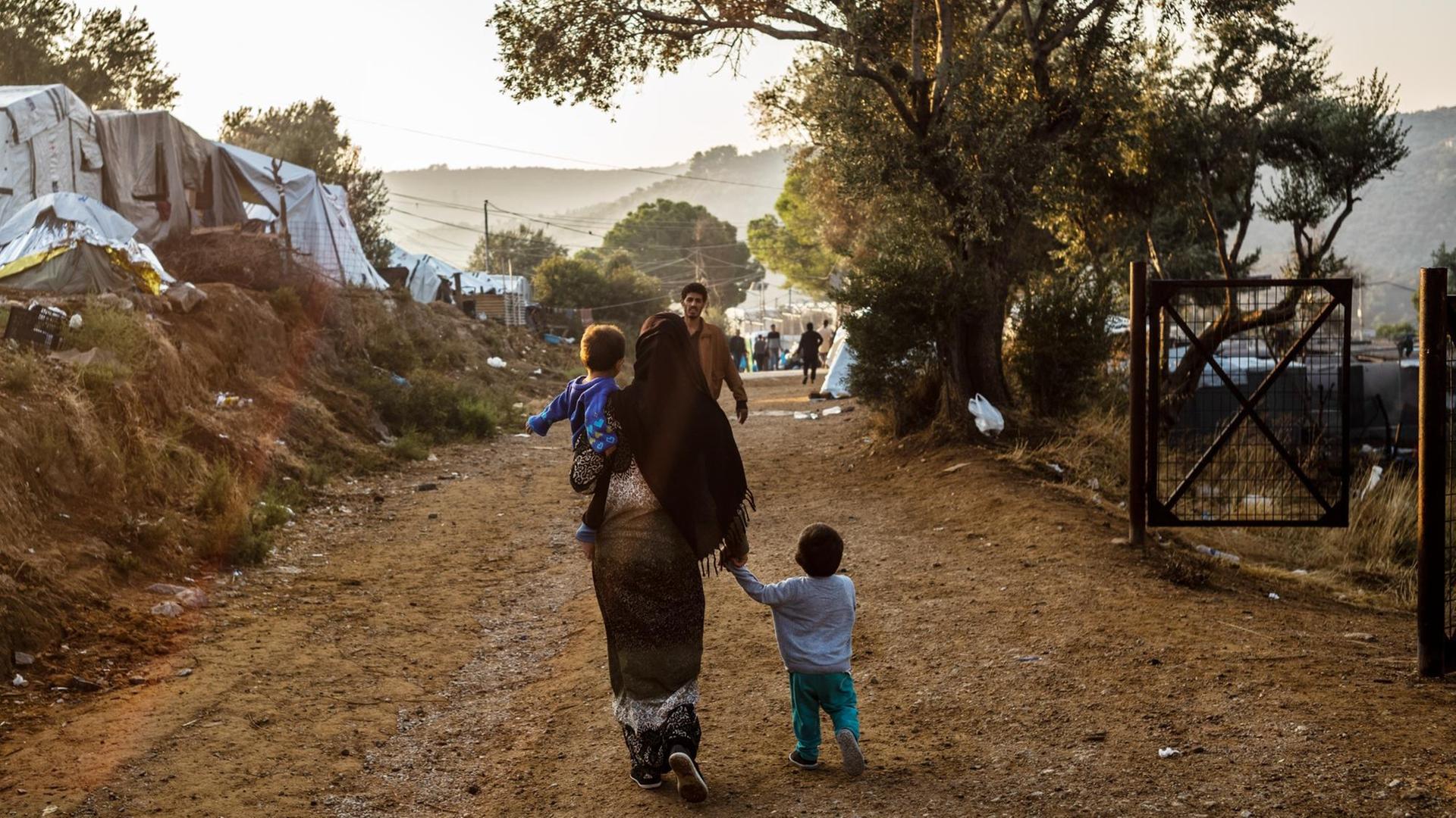 Ein Frau geht mit einem Kind auf dem Arm und einem anderen Kind an der Hand auf einem Weg im Flüchtlingslager Moria auf der griechischen Insel Lesbos, während ihr ein Mann entgegen kommt.