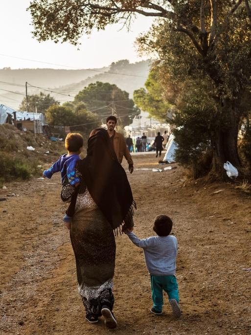 Ein Frau geht mit einem Kind auf dem Arm und einem anderen Kind an der Hand auf einem Weg im Flüchtlingslager Moria auf der griechischen Insel Lesbos, während ihr ein Mann entgegen kommt.