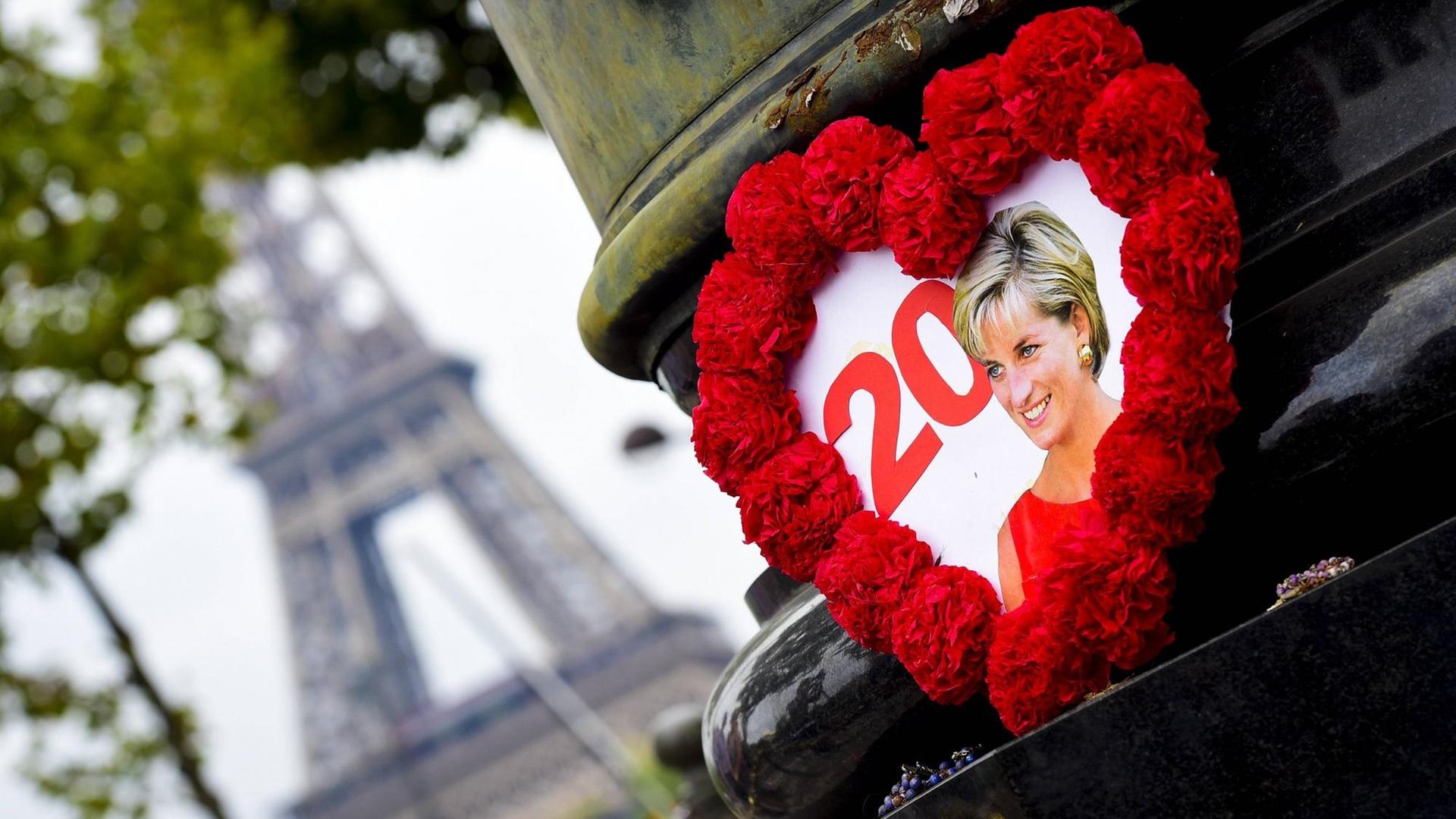 Ein gebasteltes Herz mit rotem Rahmen, in dessen Mitte ein Foto von Prinzessin Diana zu sehen ist, hängt an einer Brücke in Paris. Im Hintergrund ist der Eiffelturm zu erkennen.