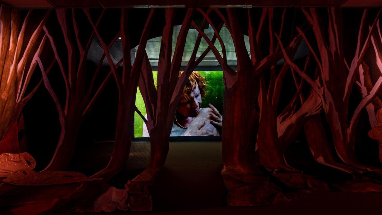Eine Installationsansicht im Hamburger Bahnhof zeigt die Komposition zweier Mixed Media Installationen von Pauline Curnier Jardin: "Qu'un Sang Impur" (2019) und "Peaux de Dame in the Hot Flashes Forest (2019). Durch eine surreal anmutende Landschaft aus roten Bäumen ist eine Leinwand mit einer älteren Frau darauf zu sehen.