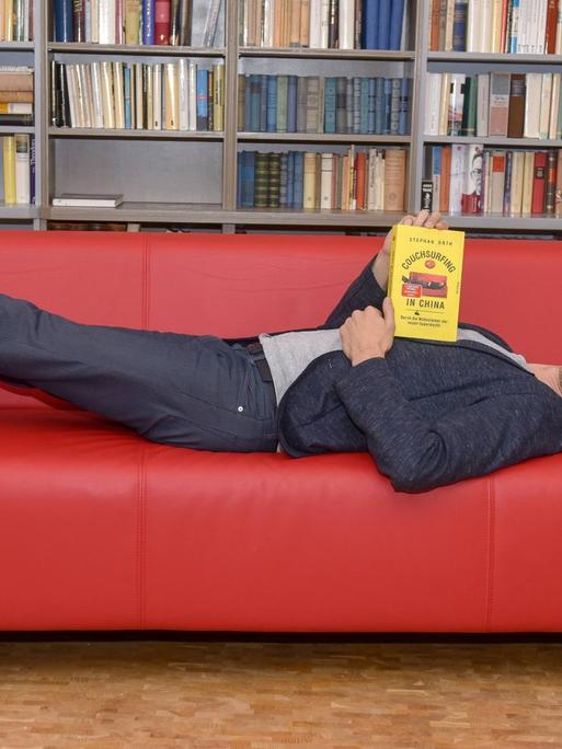 Stephan Orthliegt liegt auf einer roten Couch, einem Möbelstück, welches er auf Reisen als Couchsurfer in China in unterschiedlichen Formen vorgefunden hat. Die Erfahrungen aus dem Couchsurfing sind die Grundlage für sein neues Buch.
